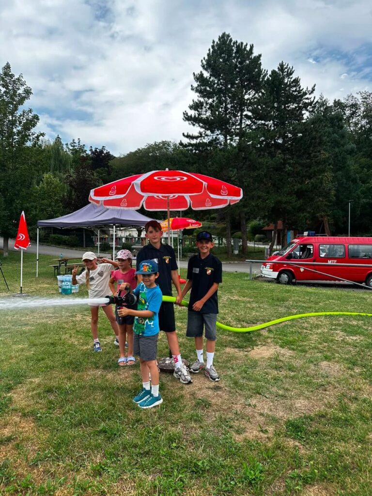 Kinder üben mit Feuerwehrausrüstung auf einer grünen Wiese vor einem roten Feuerwehrauto.