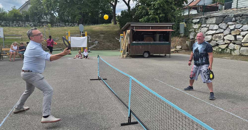 Zwei Männer spielen Pickleball auf einem Sportplatz mit einem Verkaufswagen im Hintergrund.